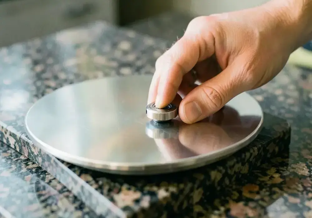 A hand checking the flatness of a polished plate with a precision dial indicator on a granite surface plate